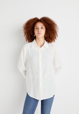 Camicia bianca a maniche lunghe con una sottile trama testurizzata, colletto a punta e orlo arrotondato. Indossata con jeans blu.