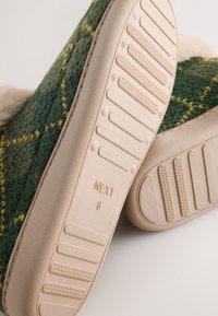 Grønne og gule ternede stofhjemmesko med beige gummisåler. Har mønstrede grebsflader og et mærke med teksten "NEXT 6."