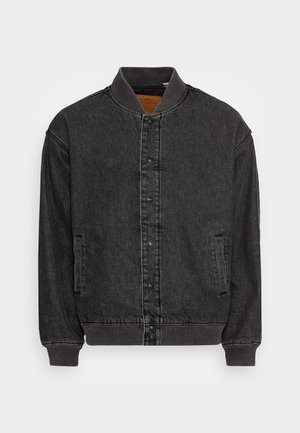 Person iført en sort bomberjakke over en orange cardigan og en stribet skjorte, lyseblå patched jeans og hvide sneakers.