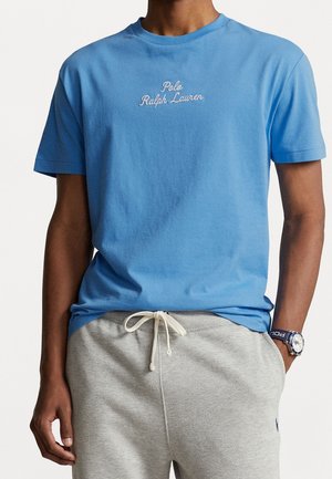 Mężczyzna w jasnobłękitnej koszulce Polo Ralph Lauren z białym logo i jasnoszarych spodniach dresowych ze sznurkiem oraz zegarkiem na lewym nadgarstku.