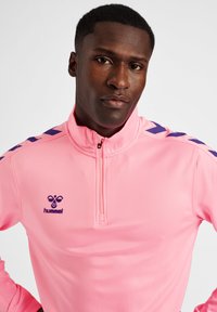 Maglia atletica rosa a metà zip realizzata in tessuto liscio, con accenti viola sulle spalle e un piccolo logo 'hummel' sul petto.