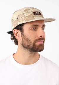 Casquette en tissu crème et beige à motifs, avec un patch logo marron, une visière plate et une sangle ajustable à l'arrière.