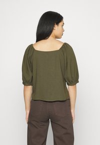 Top verde-oliva de mangas bufantes com decote quadrado e detalhes franzidos. Tecido suave e texturizado, comprimento cropped, combinado com calças castanhas de cintura alta.