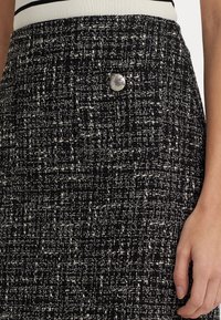 Svart och vit tweedtyg med ett strukturerat mönster, med en rund silverknappdetalj på sidan. Närbild av kjolen.