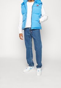 Vaaleansininen toppaliivi hupulla, valkoinen pitkähihainen paita, siniset farkut ja valkoiset tennarit sinisillä yksityiskohdilla. Liivissä on merkkilogolla varustettu merkki.