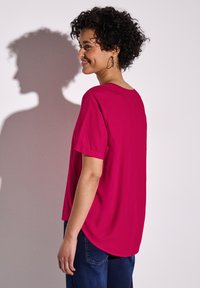 Top fucsia a maniche corte con vestibilità rilassata, orlo arrotondato e tessuto morbido. Abbinato a jeans in denim blu. Il modello mostra la vista posteriore.