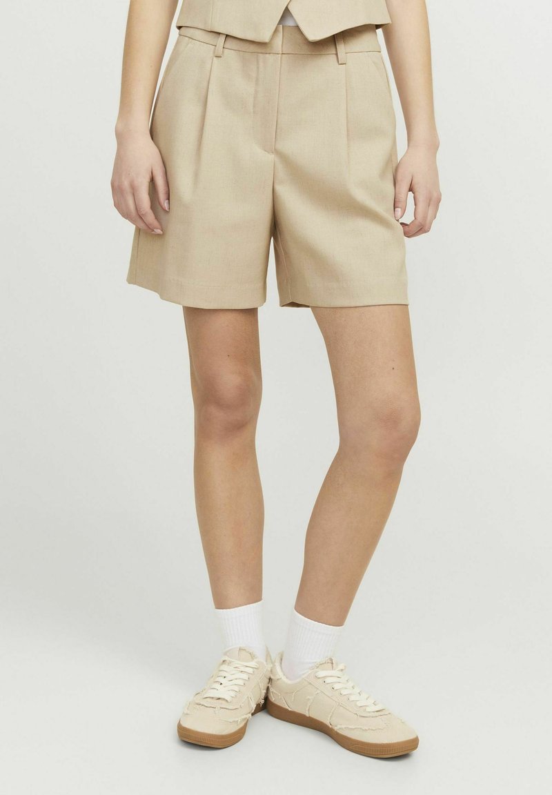 Pantalones cortos beige a medida con pliegues, de longitud media en el muslo. Combinados con zapatillas beige claras y calcetines blancos. Textura suave y líneas limpias.