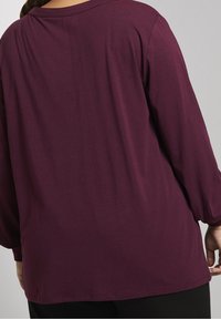 Langærmet, dyb maroon top med en afslappet pasform. Blød, glat tekstur og rund udskæring, med subtile samlinger ved ærmerne.