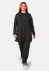 Tunic nera a coste con maniche lunghe e spacchi laterali, abbinata a pantaloni larghi coordinati. L'outfit presenta una vestibilità comoda ed è abbinato a sneakers.