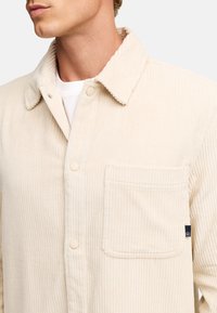 Camicia in corduroy beige con colletto a pieghe, bottoni frontali e tasca. Tessuto testurizzato con righe verticali e etichetta con logo sul lato.