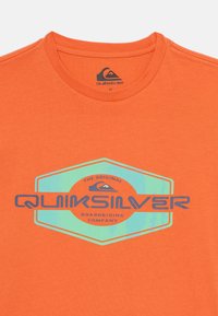 Oranje katoenen T-shirt met een hexagonaal grafisch ontwerp waarop de tekst "Quiksilver" in het blauw staat, met een verloop van groene achtergrond. Ronde halslijn.