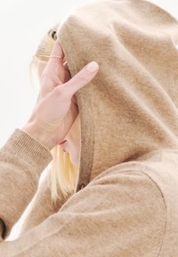 Sudadera beige hecha de un tejido suave y texturizado, que presenta una capucha con cordón y puños acanalados. La mano del portador sostiene la capucha parcialmente levantada.