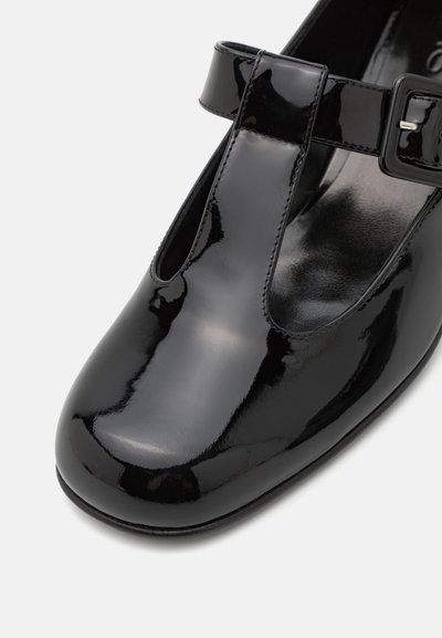 Zapato de charol negro con punta redondeada, correa en T y detalle de hebilla. La textura suave y el acabado brillante realzan su apariencia elegante.