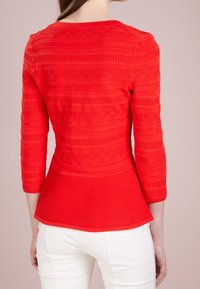 Femme portant un haut rouge texturé à manches longues cintré avec un ourlet à volant, associé à un pantalon blanc, vue de dos.