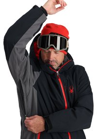 Black and gray ski jacket with red zipper, logo, and hood. Features a zippered pocket and textured fabric, designed for winter sports.