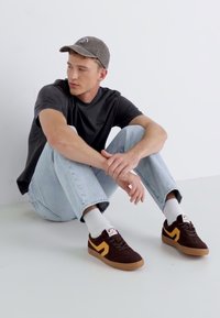 Dunkelbraune Wildledersneaker mit gelben Akzenten, weiße Socken, hellblaue Jeans und ein schwarzes T-Shirt, getragen von einem sitzenden Modell.
