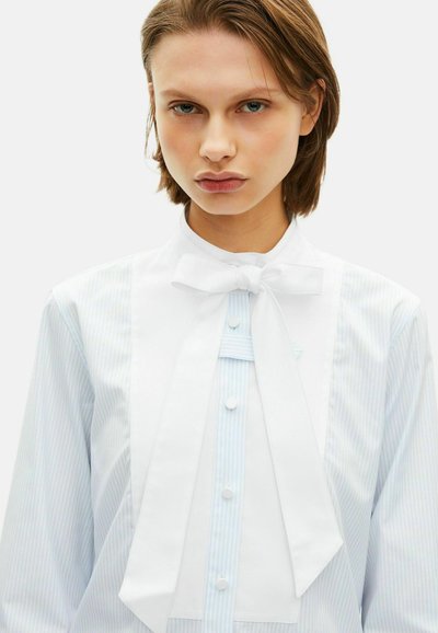White blouse with a bow tie, featuring light blue vertical stripes. Button-up front with plain white bib and collar at neck.