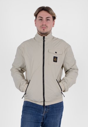 Giacca leggera beige con colletto alto, zip frontale e tasca sul petto con bottoncino nero e patch con logo.