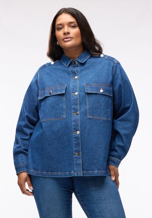 Donna che indossa una giacca di jeans blu abbottonata con due tasche sul petto e jeans coordinati su uno sfondo semplice.