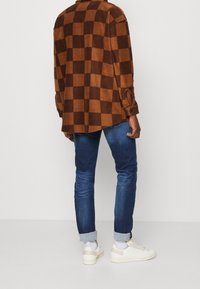 Chemise en flanelle à carreaux bruns et beiges, texture douce, coupe décontractée, associée à un jean en denim bleu et des baskets blanches avec des accents beiges.