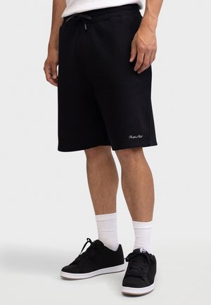 Άνδρας που φοράει μαύρα σορτς με κορδόνι, άσπρες κάλτσες και μαύρα αθλητικά παπούτσια, στέκεται μπροστά σε ανοιχτό γκρι φόντο.