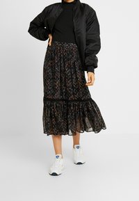 Jupe florale noire avec un ourlet volanté, texture de tulle superposée, associée à une veste bomber noire et des baskets blanches avec des accents bleus.
