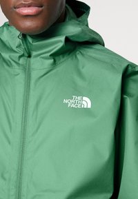 Gros plan d'une personne portant une veste verte North Face avec une fermeture éclair à l'avant et une capuche, montrant le logo de la marque sur la poitrine.