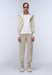 Beige huppari logolla, kerrostettuna beiget vetoketjullinen liivi; yhdistettynä samansävyisiin housuihin ja vaaleanbeigeihin korkeatossuihin.