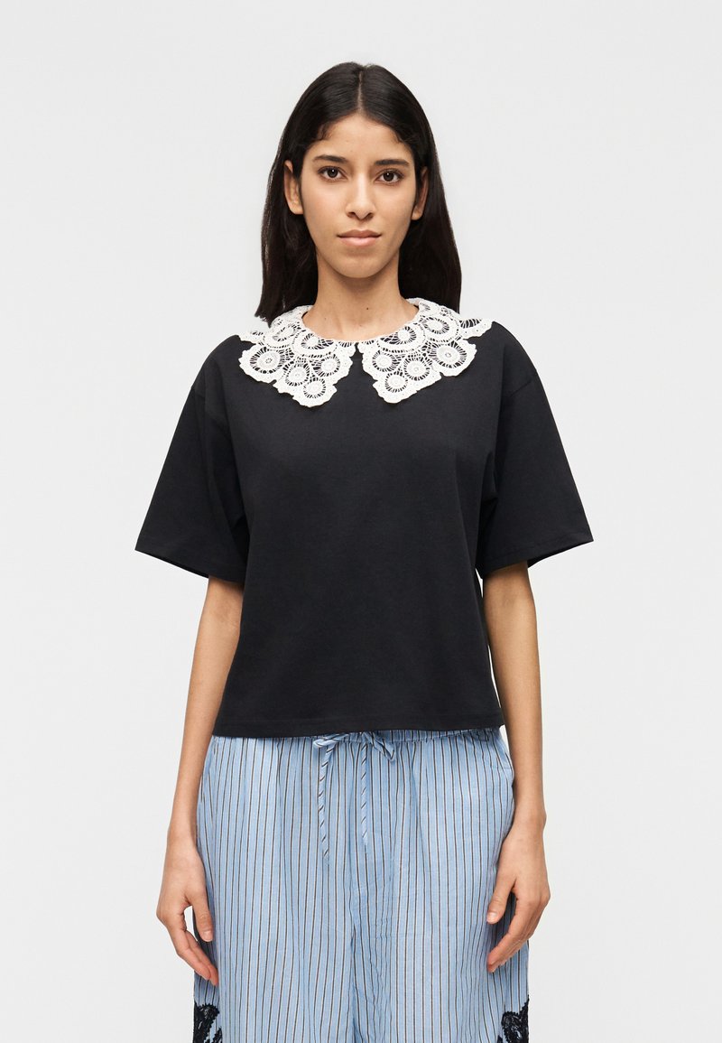 Jeune femme portant un haut noir à manches courtes avec un col en dentelle blanche et un pantalon bleu clair à rayures avec une ceinture à cordon.