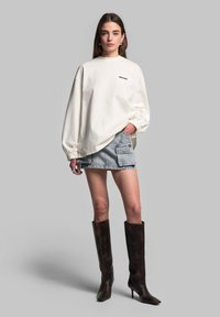 Witte oversized sweatshirt met verlaagde schouders, gecombineerd met een denim mini-jurk met een zijzak, en bruine kniehoge haklaarzen.