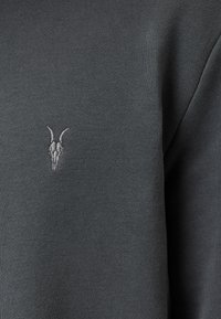 Suéter de punto gris oscuro con un pequeño emblema de cráneo de cabra gris claro en la zona izquierda del pecho. Textura suave, diseño ajustado.