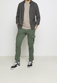 Chemise grise boutonnée portée sur un t-shirt beige, pantalon cargo vert avec poches latérales, et baskets noires avec des accents blancs.