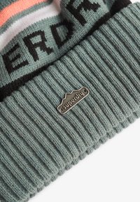Dziergany beanie w przytłumionym odcieniu teal z prążkowaną teksturą. Posiada pasek w paski w kolorze pomarańczowym i białym oraz metalową naszywkę z wytłoczonym napisem "Superdry."