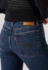 Calças de ganga azul escuro, de cintura alta, com um emblema de pele da marca e costura dourada distinta em padrão curvo nos bolsos.