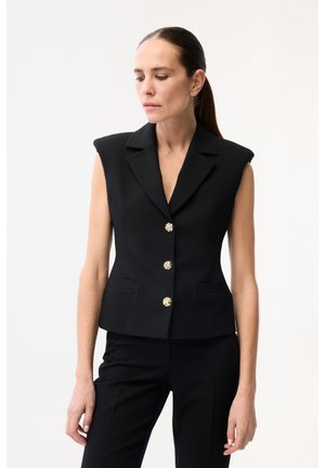 Vrouw met donker haar in een strakke paardenstaart, gekleed in een mouwloos zwart blazer met drie sierlijke gouden knopen en bijpassende zwarte broek.