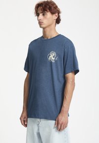 Camiseta de algodón azul con mangas cortas, que presenta un estampado gráfico de color claro en la parte izquierda del pecho. Ajuste relajado, cuello redondo.