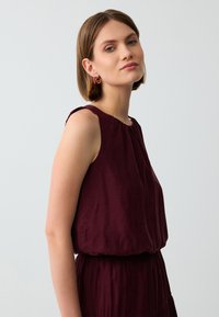 Top senza maniche borgogna con una texture morbida e scollatura plissettata, caratterizzato da una vestibilità ampia e delicati dettagli sulle spalle.