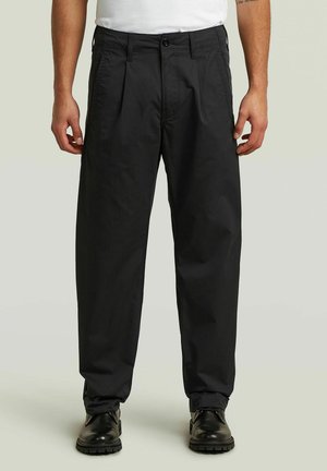 Homme portant un pantalon noir plissé ample, des chaussures en cuir noires et une chemise blanche sur un fond uni clair.