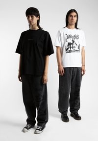 Due modelli sono in piedi uno accanto all'altro. Uno indossa una camicia nera oversize e pantaloni larghi scuri. L'altro indossa una maglietta bianca con grafica e pantaloni grigi.