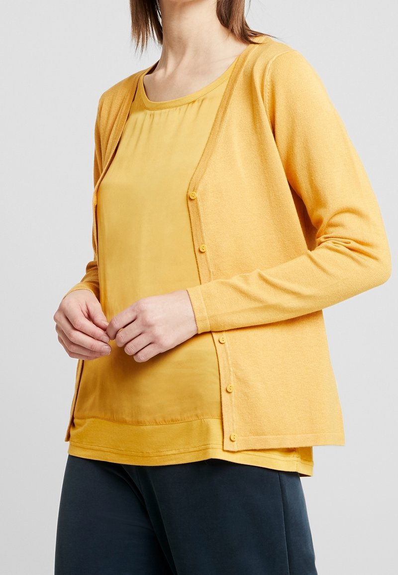 Femme portant un cardigan moutarde boutonné sur un chemisier assorti, associé à un pantalon foncé, les mains croisées devant elle.