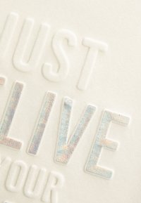 Tkanina w kremowym kolorze z wytłoczonym napisem "JUST LIVE" i lśniącymi, wielokolorowymi akcentami na literach, co nadaje tekstuuralny wygląd.
