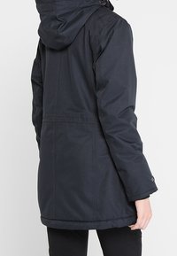 Mörk, huva jacka med en texturerad tyg, som har en rak skärning, långa ärmar och en fåll som når mitt på låret.