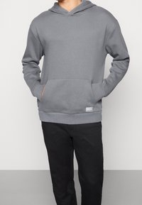 Hoodie in pile grigio con tasca a marsupio, polsini e orlo ribattuti, e cappuccio foderato. Indossato con pantaloni neri.