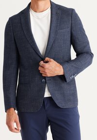 Muž zapíná knoflík na tmavě modrém texturovaném saku nošeném přes bílou košili s tmavě modrými kalhotami, ukazuje prsteny na prstech a tenký náhrdelník.