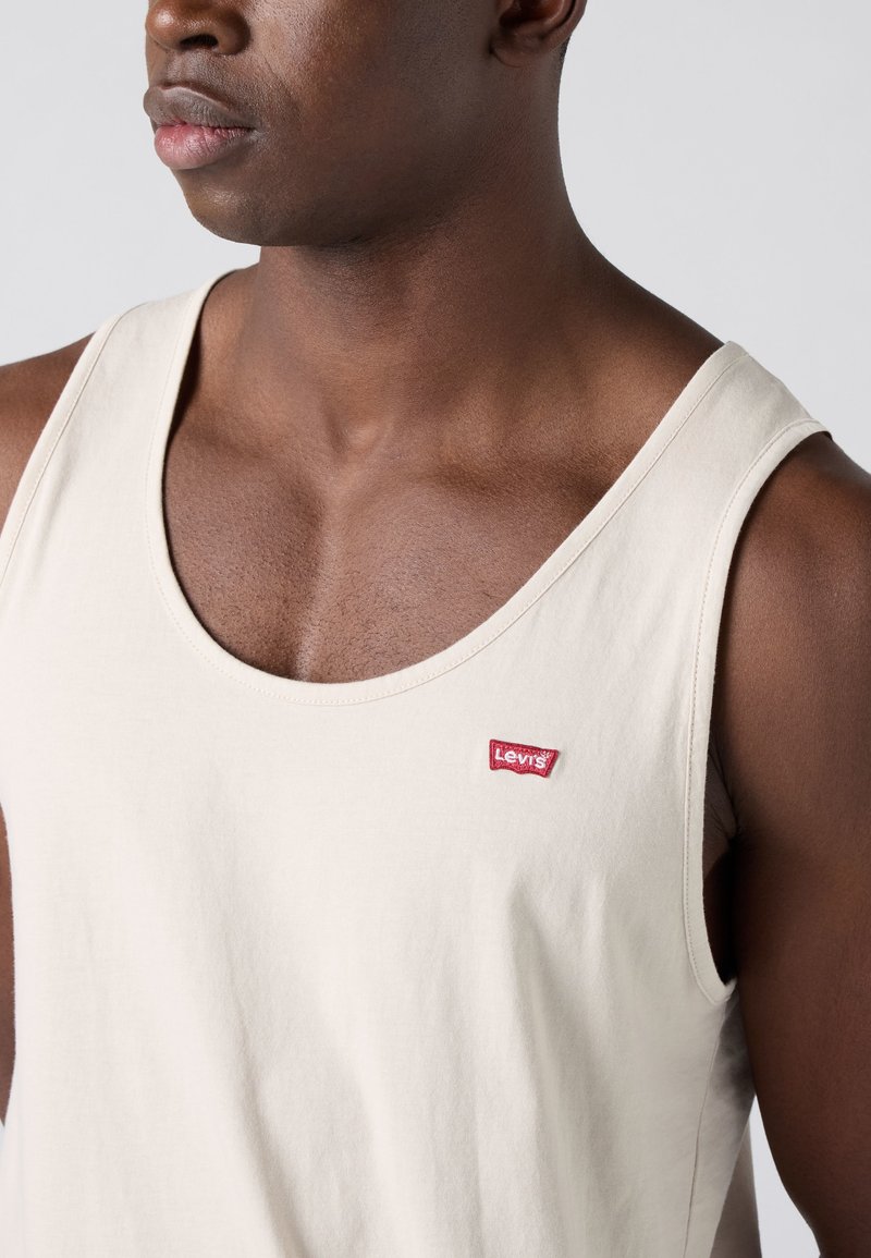 Gros plan sur un homme à la peau foncée portant un débardeur Levi’s beige clair, montrant la poitrine et les épaules sur un fond uni.