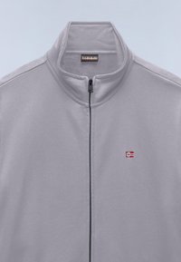 Jasnoszara bluza polarowa na zamek błyskawiczny z kołnierzem stojącym oraz małym czerwono-biało-niebieskim logo na lewej piersi.