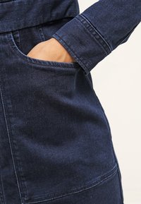 Laivastonsininen farkkutakki, jossa on näkyvällä ompelulla varustettu tasku, teksturoitu kangas ja istuva malli. Hihassa näkyy kalvosimen yksityiskohta.