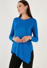 Top blu, a maniche lunghe con scollatura rotonda e orlo asimmetrico. Realizzato in tessuto morbido, presenta uno stile aderente. Abbinato a pantaloni neri.