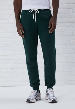 Pantalons de survêtement verts en tissu doux avec cordon blanc, design fuselé et poignets élastiques, associés à des chaussures de sport argentées.