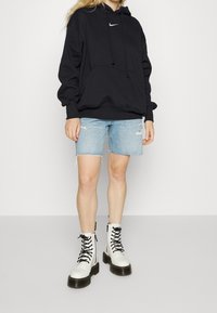 Svart huvtröja med frontficka, ljusblå slitna jeansshorts och vita snörstövlar med tjocka svarta sulor.